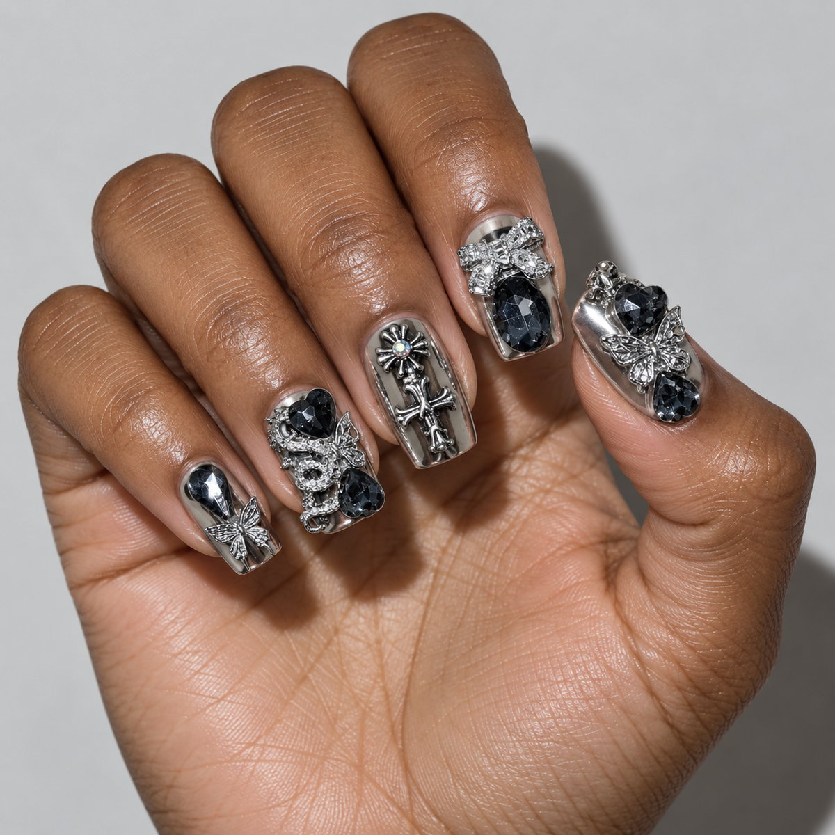 A close-up of a hand showcasing a gothic-style nail art design. The nails feature silver metallic plates, intricate filigree, small silver crosses, and dark blue faceted gemstones.