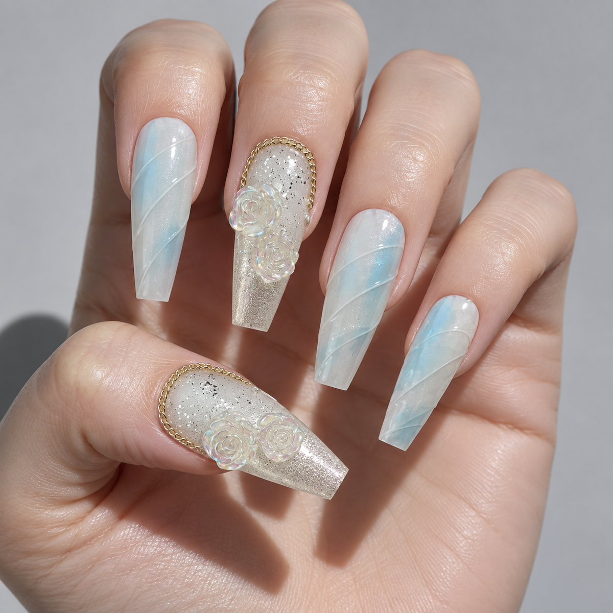 A close-up of a hand showcasing a set of long coffin-shaped press-on nails. The nails feature a light blue and white diagonal swirl pattern on some, a glittery white base with 3D floral rosettes on the accent nails, and a thin gold metallic border.
