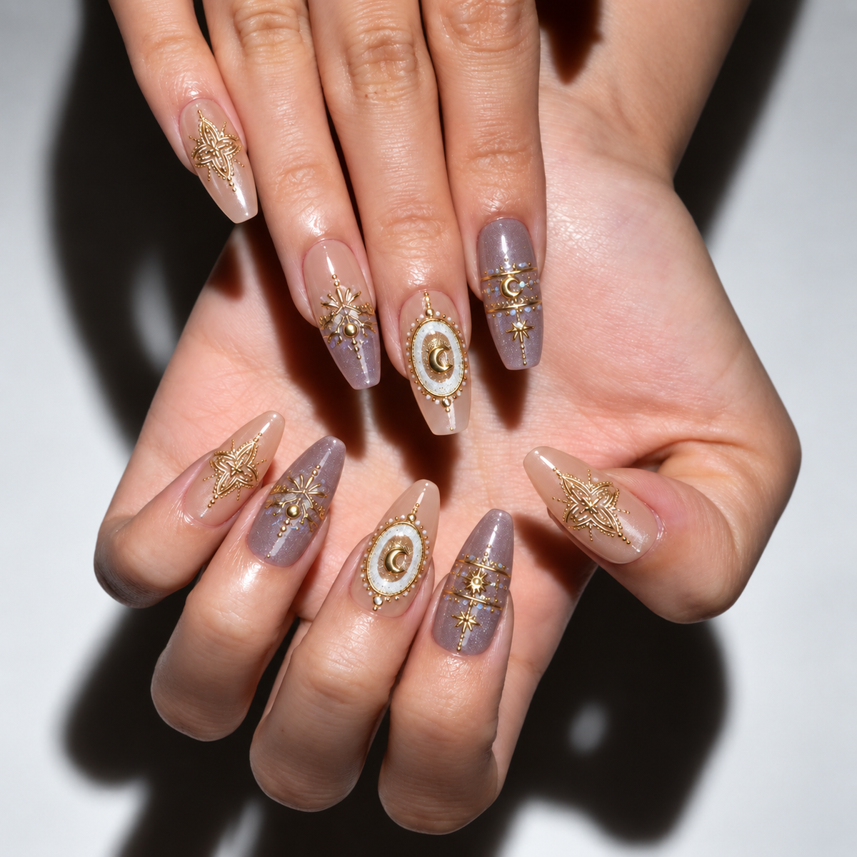 A close-up of a person's hands showcasing a detailed coffin-shaped nail art design. The nails feature a sheer lavender-purple base with metallic gold starburst patterns, filigree, and circular medallion designs with white and gold accents.