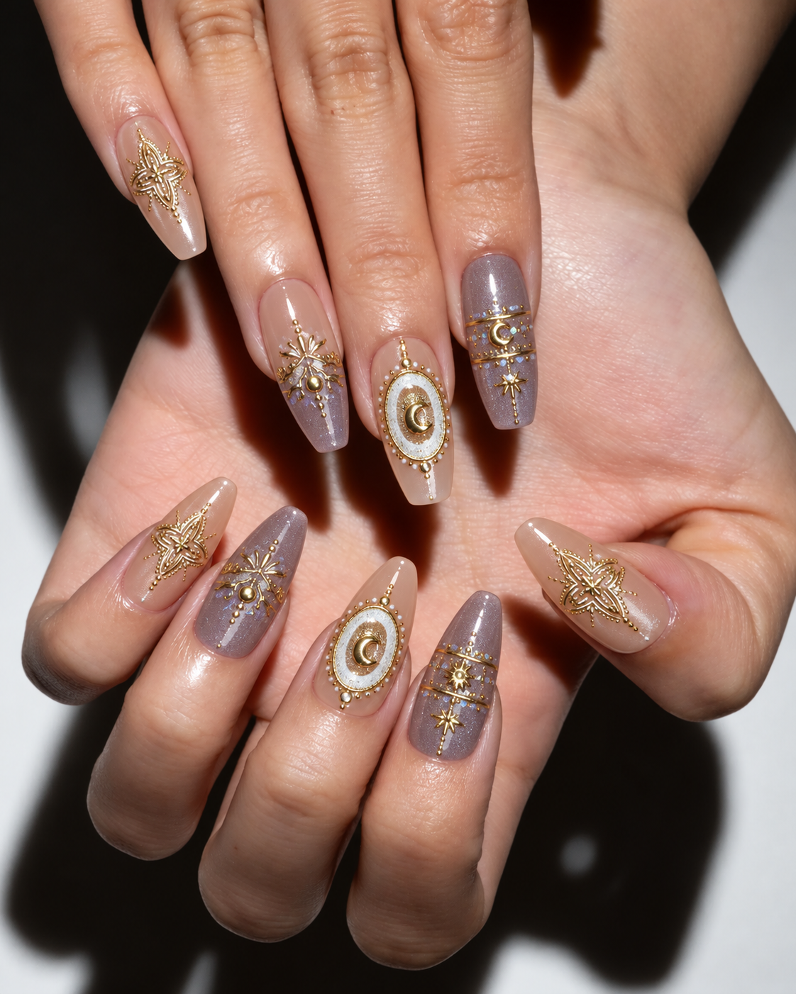 Close-up of hands showcasing a detailed coffin-shaped nail art design. The manicure features a glossy nude base with lavender-gray tips, gold celestial star patterns, and central oval designs with white and gold details.