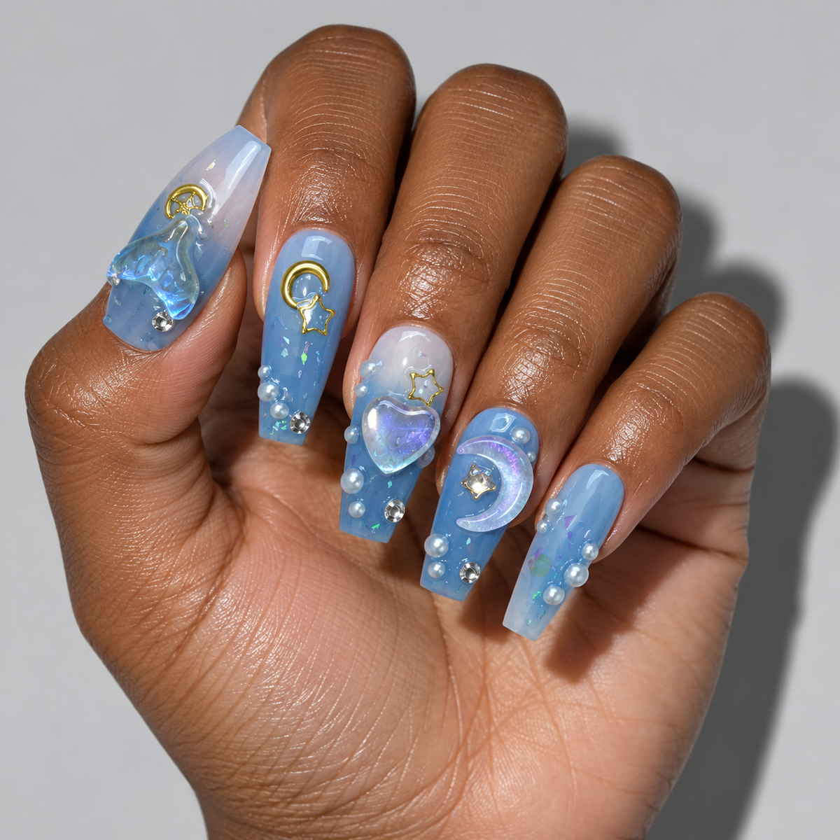A hand with long, decorated press-on nails featuring a celestial theme in shades of light blue, white, and gold. The nails are embellished with 3D glass charms, rhinestones, and small pearl accents against a neutral gray background.