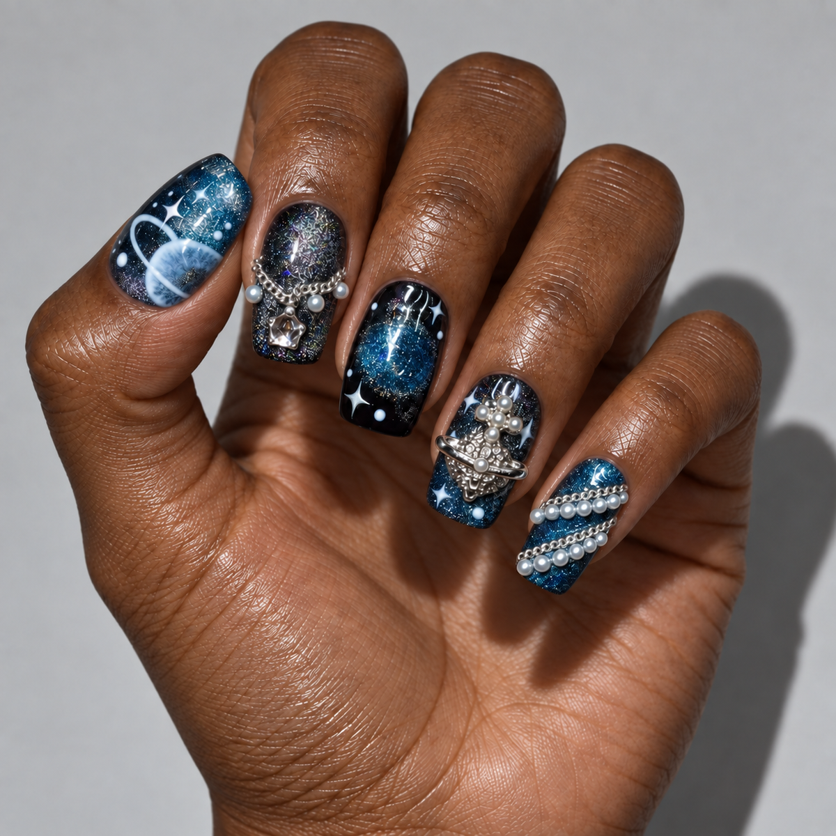 A close-up of a hand showcasing a detailed galaxy-themed nail art design. The manicure features a mix of deep blue and black glittery galaxy patterns, silver rhinestone skull motifs, white pearl accents, and star details on each fingernail, set against a neutral gray background.