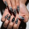 A close-up of hands showcasing long, sculpted stiletto-shaped press-on nails with a gothic silver and black design, featuring rhinestone embellishments and metallic details.