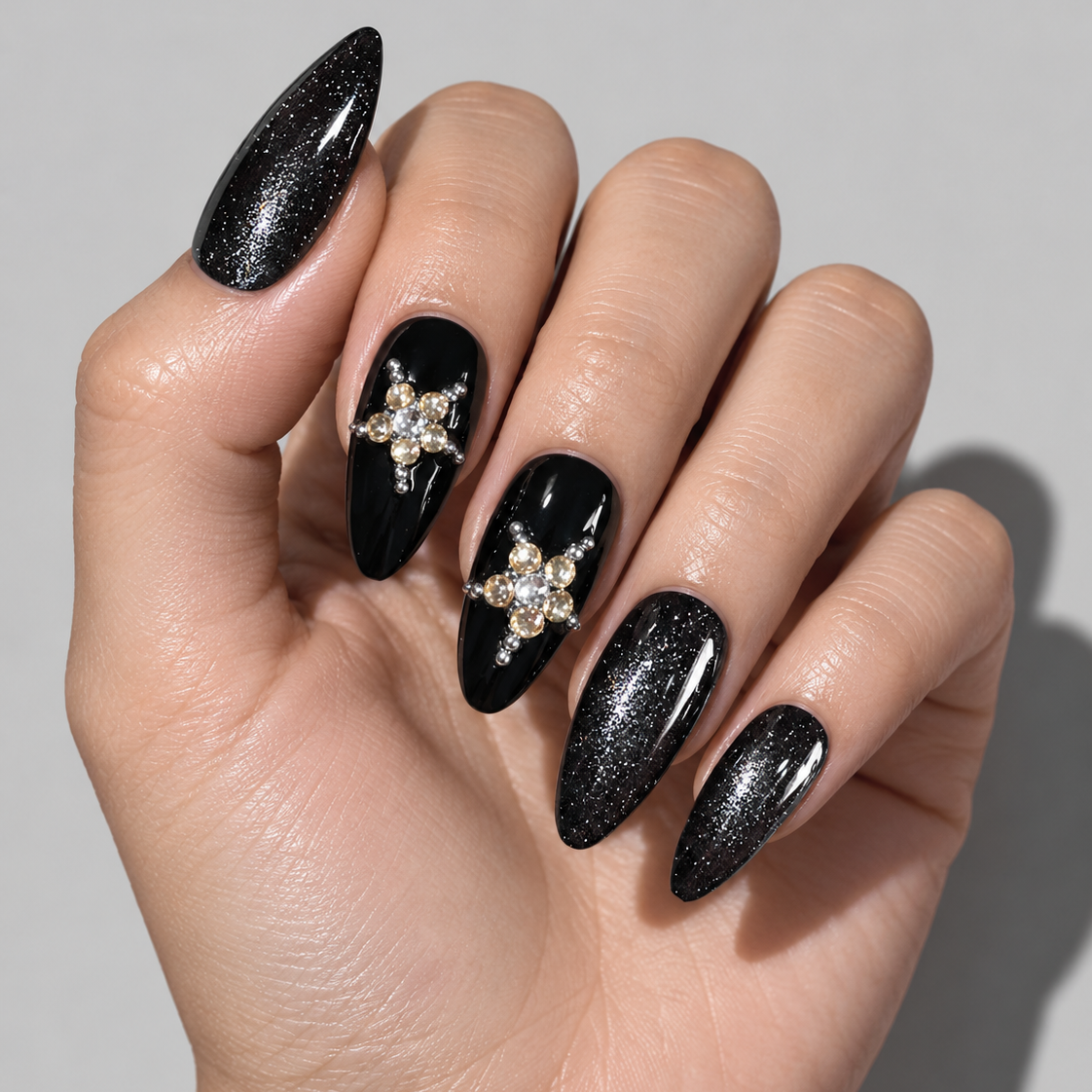 A close-up of a hand with long stiletto-shaped press-on nails. The nails are primarily black with one glittery black nail, one glossy black nail with a gold and clear rhinestone star design, and one glittery black nail.