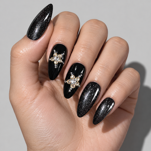 A close-up of a hand with long stiletto-shaped press-on nails. The nails are primarily black with one glittery black nail, one glossy black nail with a gold and clear rhinestone star design, and one glittery black nail.