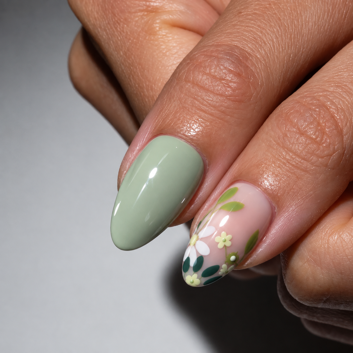 Close-up of a hand showing manicured fingernails, with one nail painted a soft sage green and the other decorated with white flowers and green leaves on a pale pink base.