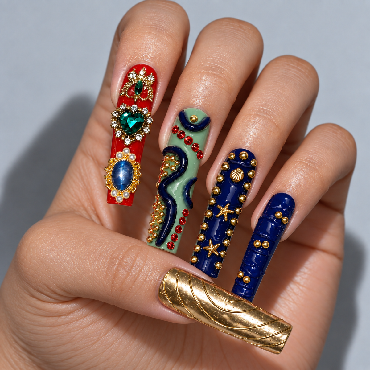 A close-up of a hand showcasing a set of long, ornate coffin-shaped press-on nails with intricate designs. The nails feature a mix of colors and patterns: one is red with jewel-like embellishments, one is mint green with gold and red accents, one is dark blue with gold stars and shells, and the thumb is a textured metallic gold.