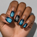 A close-up of a hand showcasing a detailed nail art design. The nails feature a mix of deep teal blue and black backgrounds decorated with metallic gold celestial motifs, including a bow and arrow, a snowflake, and stars.