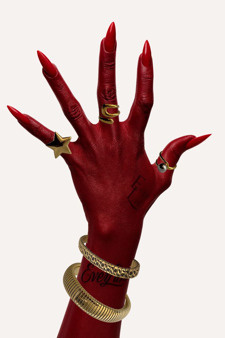 A hand with long red nails and red-painted skin, wearing multiple gold rings and thick gold bracelets, set against a plain off-white background.